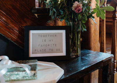 A floral arrangement in soft pinks and greens sits by a wooden staircase next to a framed sign reading “Together is my favorite place to be” and a small wrapped gift.