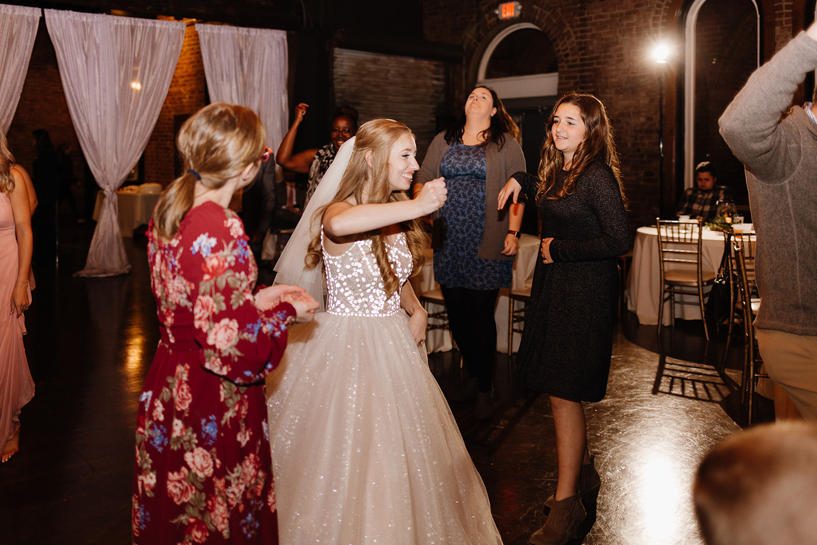 A close-up of wedding bouquets in glass vases on a black marble table, featuring blush and white roses, eucalyptus, and thistle accents, with a softly lit card box in the background.