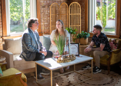 Three people sit and chat in a sunlit lounge area with cozy seating, greenery, and exposed brick walls.