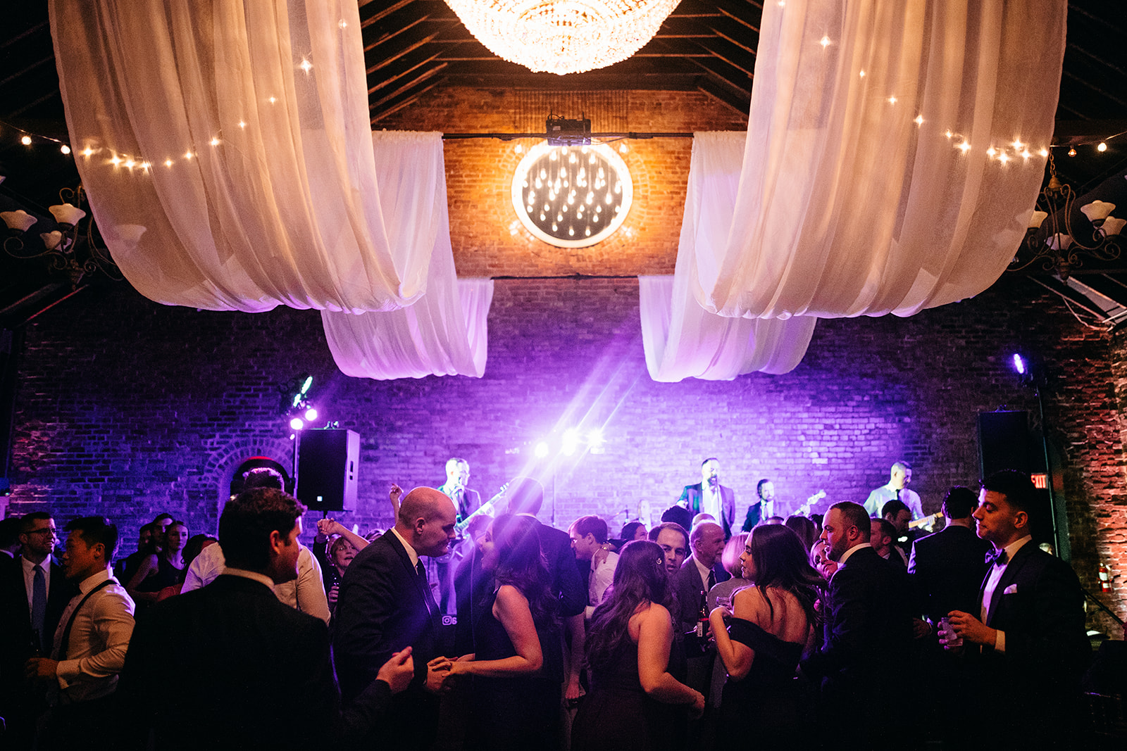 A close-up of wedding bouquets in glass vases on a black marble table, featuring blush and white roses, eucalyptus, and thistle accents, with a softly lit card box in the background.