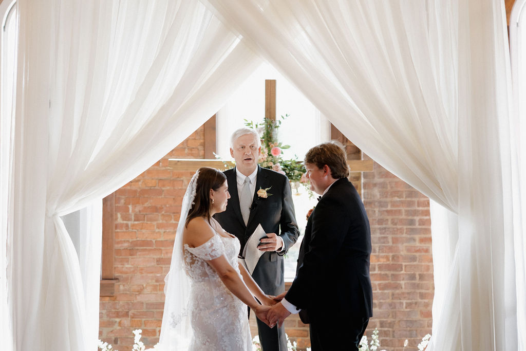 A man in a navy suit and a woman in a floral dress stand smiling together against an exposed brick wall.